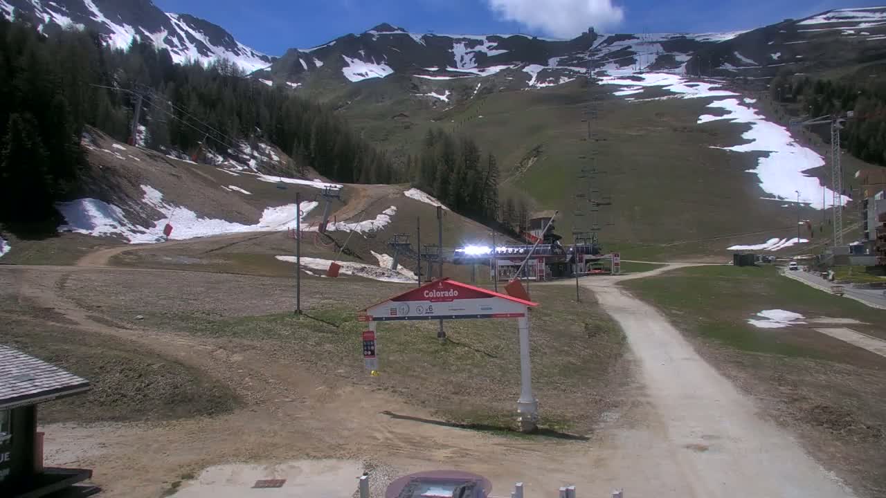 A mostly sunny view of a ski resort in spring, showing grassy slopes with patches of remaining snow, ski lifts, and a dirt road leading to a building.