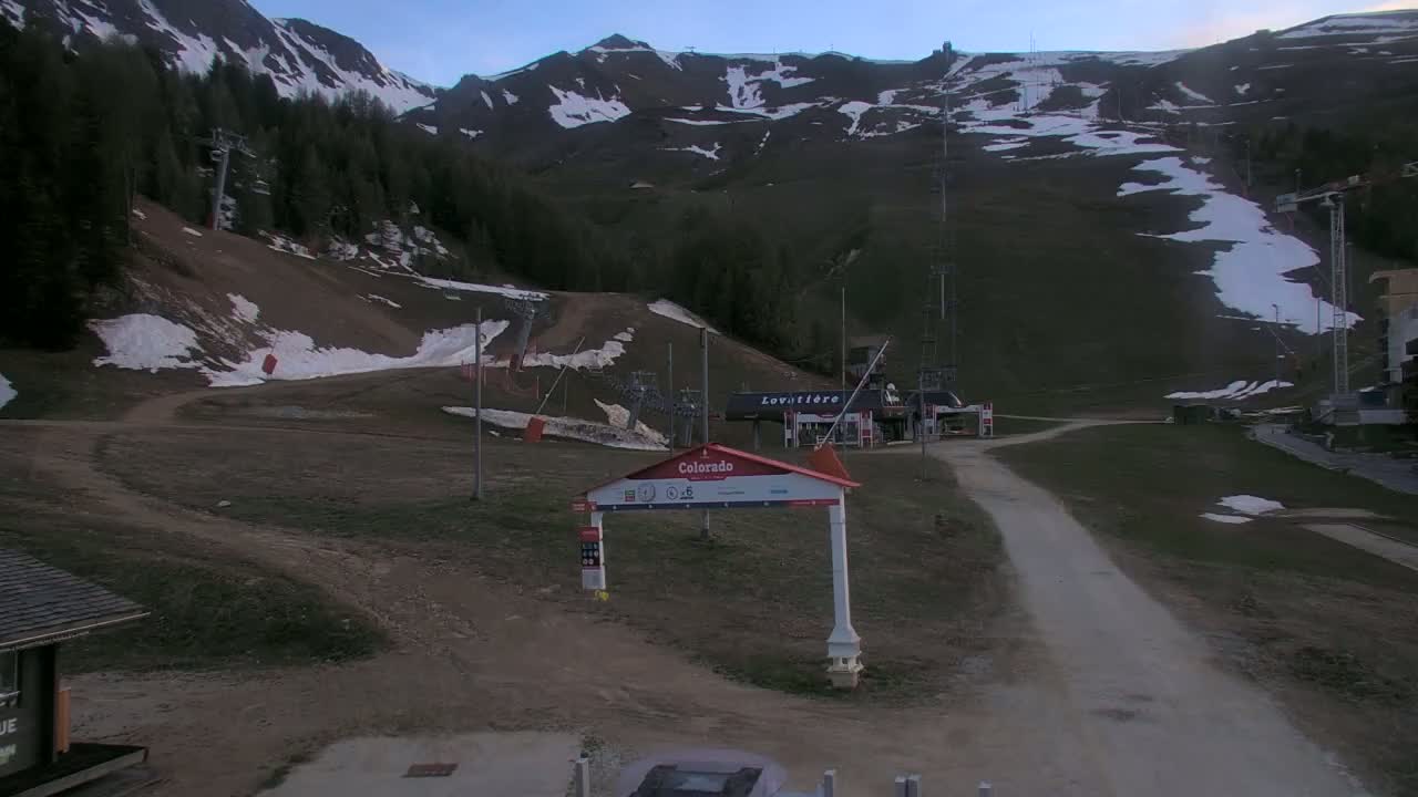 A mountainside ski resort with patches of remaining snow under a twilight sky shows a dirt path leading to a lift station.
