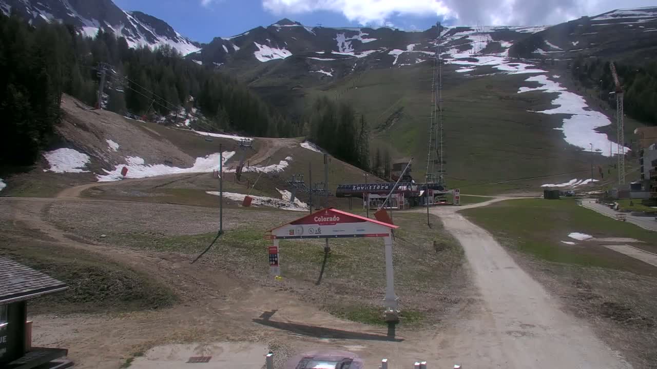 A mostly clear, sunny day shows a mountainside ski resort with patchy snow, dirt roads, and ski lifts.