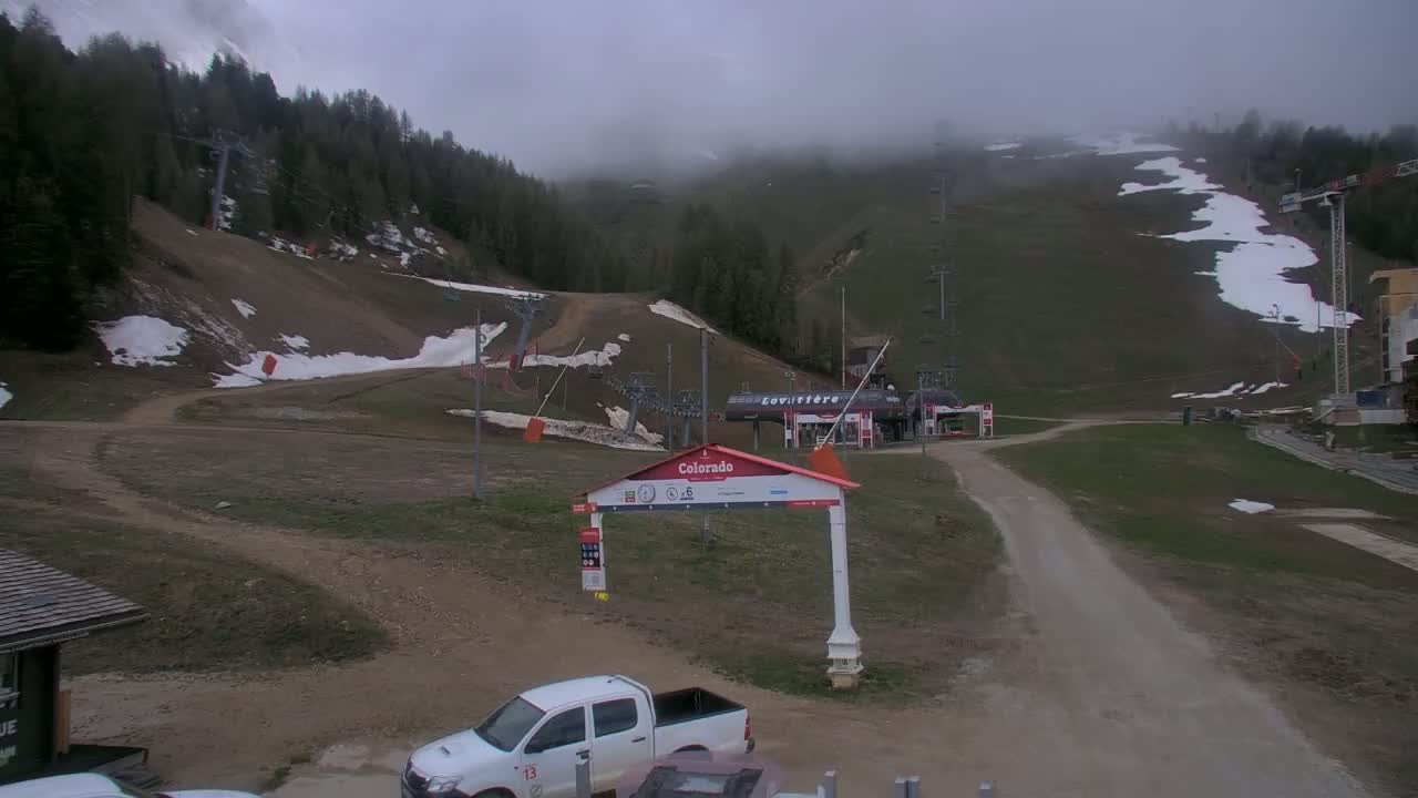 A mostly cloudy day at a ski resort shows patchy snow on the slopes, with a dirt road leading to a lift station, and a white pickup truck parked nearby.