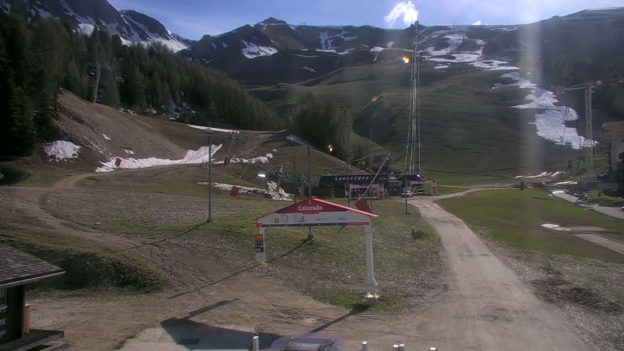 A sunny day at a ski resort shows mostly green grassy hills with patches of remaining snow, ski lifts, and a dirt road leading to a building.