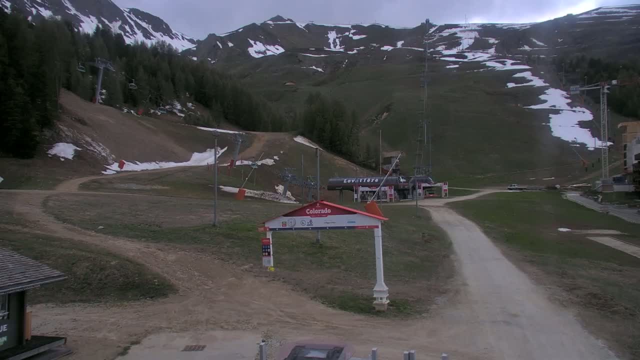 A mostly dirt road leads to a ski lift base area with some remaining snow patches on the surrounding grassy mountains under an overcast sky.