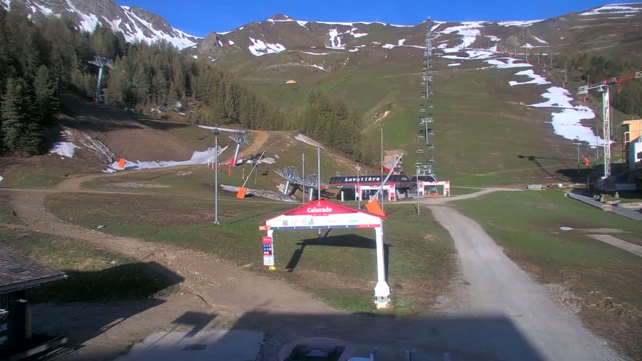 A sunny day at a ski resort shows mostly green grassy slopes with patches of remaining snow, several chairlifts, and a base area with buildings.