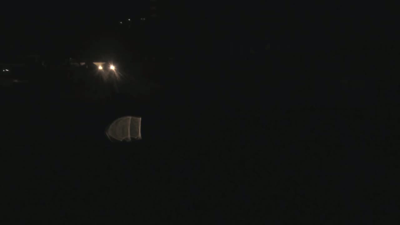 A dark nighttime scene shows a faint reflection of headlights on a dark surface, with indistinct shapes and lights in the distance.