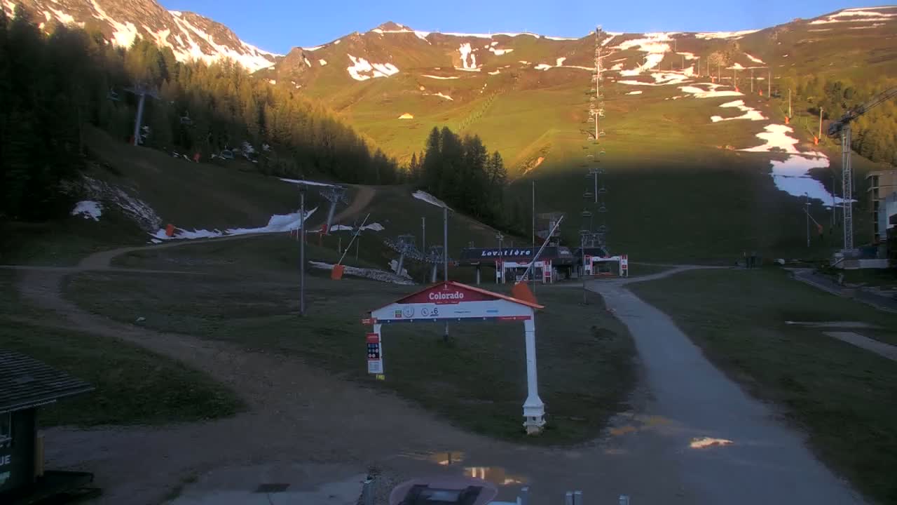 A ski resort with grassy slopes, remnants of snow, and chairlifts, illuminated by the soft light of sunrise or sunset.