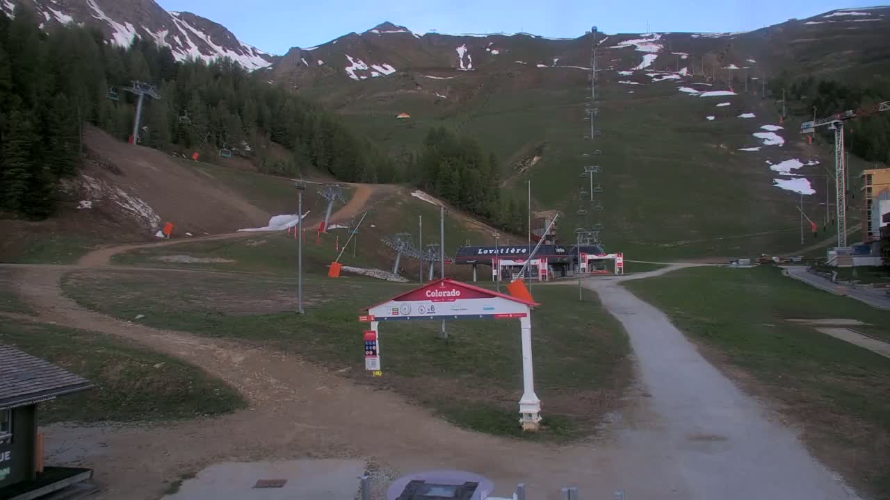 A mostly clear, cool-looking day at a ski resort, showing grassy areas, ski lifts, and a mountainside with patches of snow.
