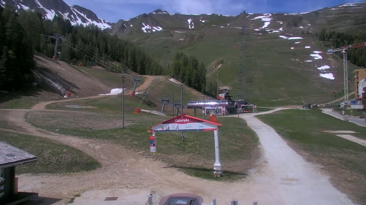A mostly sunny day at a ski resort shows a grassy mountainside with remnants of snow, ski lifts, and a dirt road leading to a lift station.