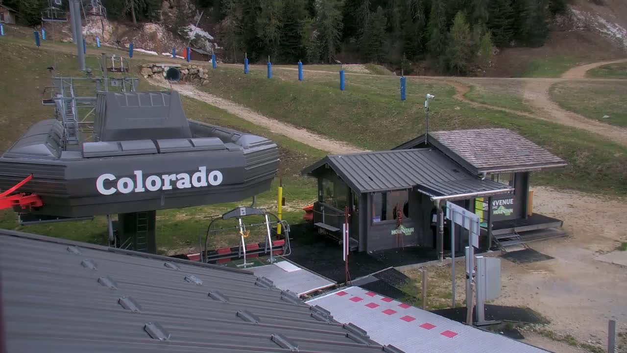 A chairlift terminal labeled "Colorado" is situated on a grassy hill beside a small building, under a partly sunny sky.