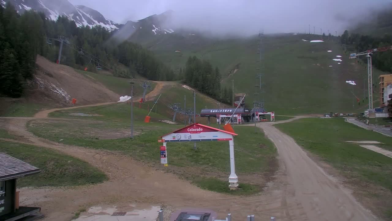 A mostly cloudy day reveals a ski resort with chairlifts and a dirt road winding through grassy areas, with some remaining snow patches visible on the mountainside.