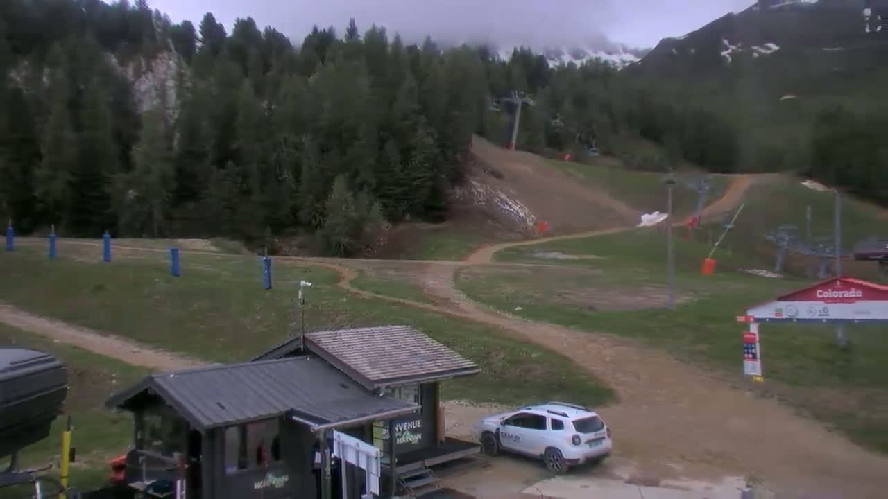 A mostly cloudy day at a ski resort shows a grassy area with dirt paths, a small building, a white SUV, and a ski lift in the background.