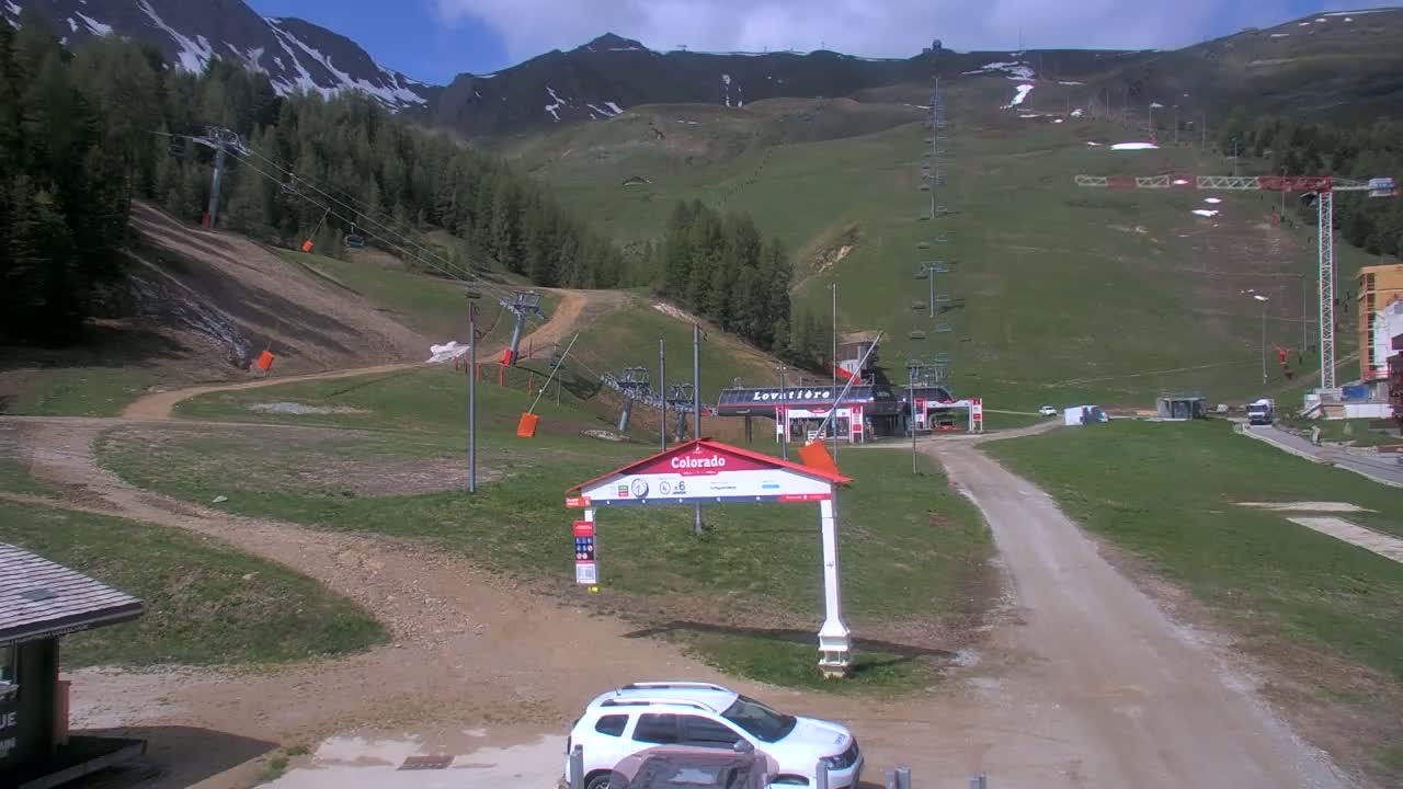 A mostly sunny day at a ski resort shows grassy hills, chairlifts, and a small white car parked near a dirt road leading to a ski lodge, with some snow still visible on the distant mountains.