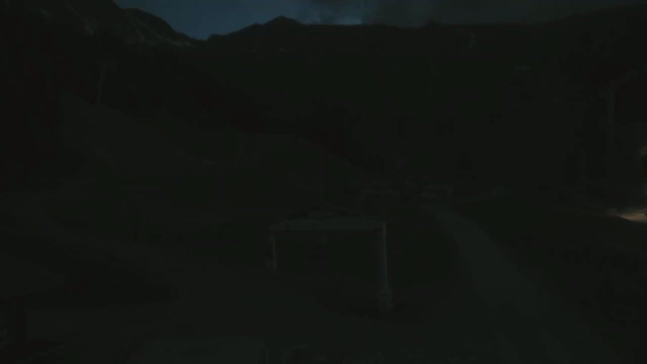 A dark, nighttime scene shows a dimly lit structure and a road leading toward mountains under a dark, cloudy sky.