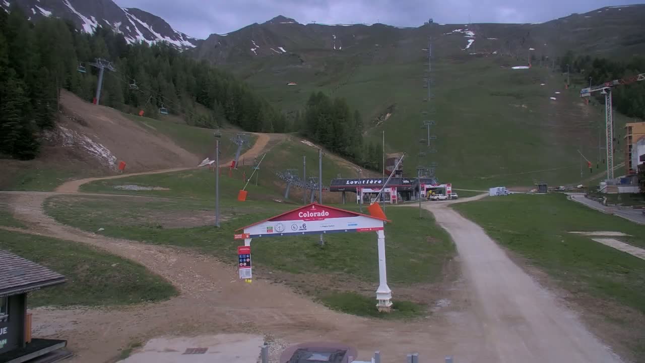 A mostly cloudy day at a ski resort in the mountains shows a dirt road leading to a ski lift base area and a mountain with patchy snow.