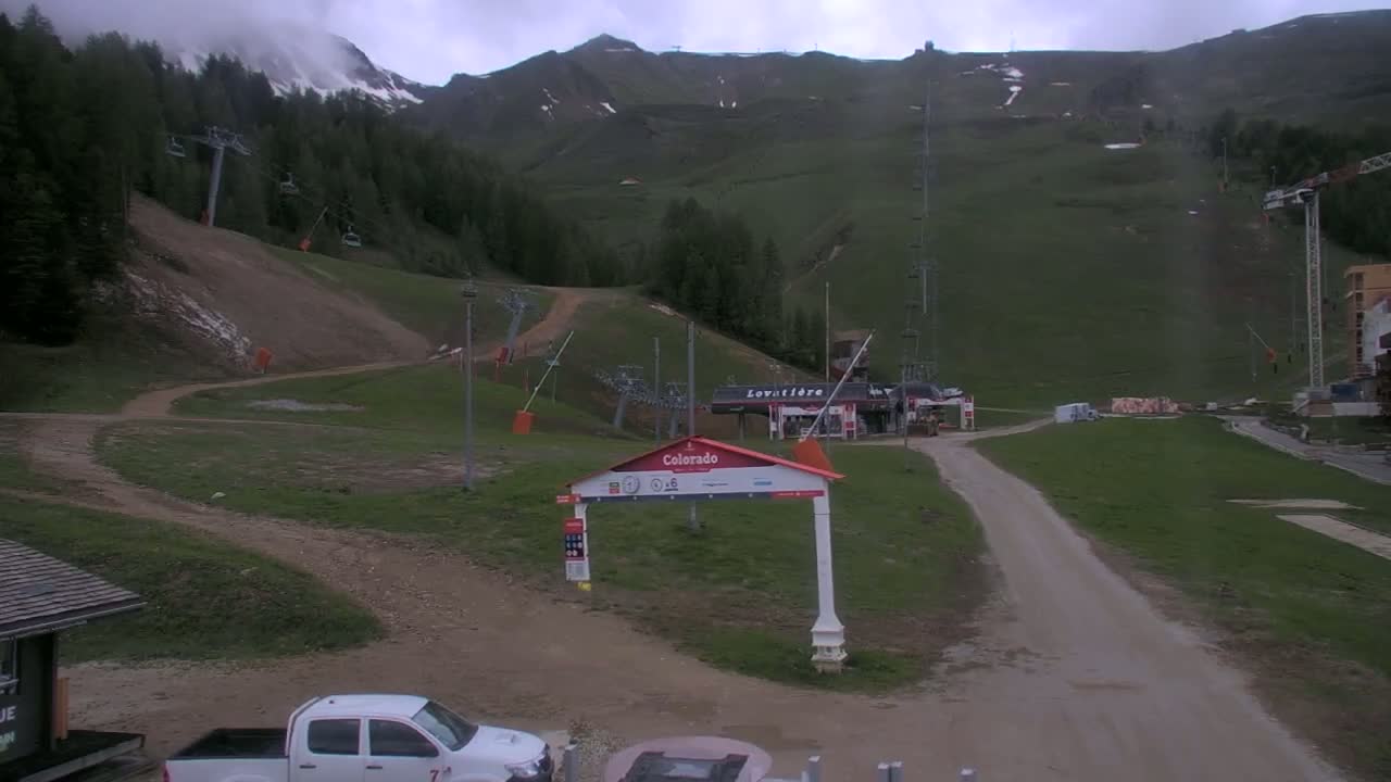 A mostly cloudy day at a mountain ski resort shows a dirt road leading to a lift station, with ski lifts and some construction visible on the grassy slopes.