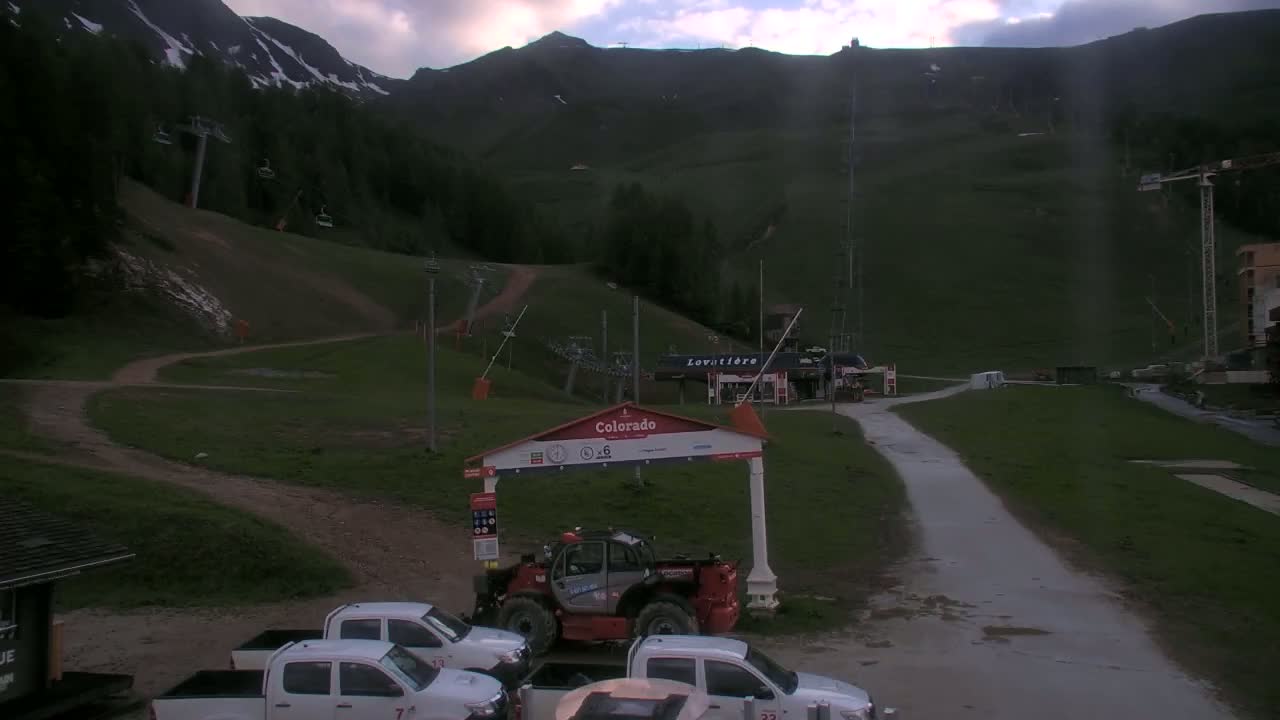 A mountainside ski resort with several chairlifts, some parked vehicles, and overcast skies.