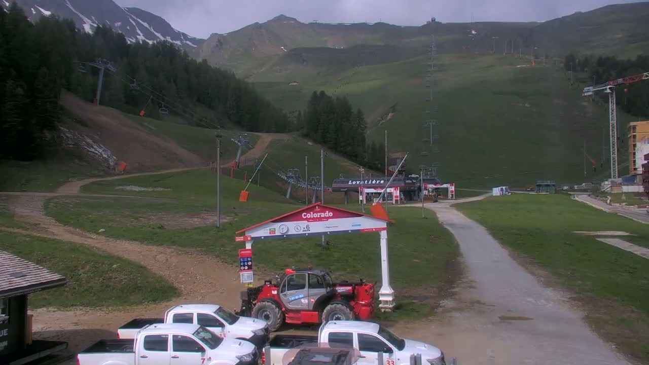 A mountain ski resort on an overcast day shows several chairlifts, a dirt road, parked trucks, and construction equipment.
