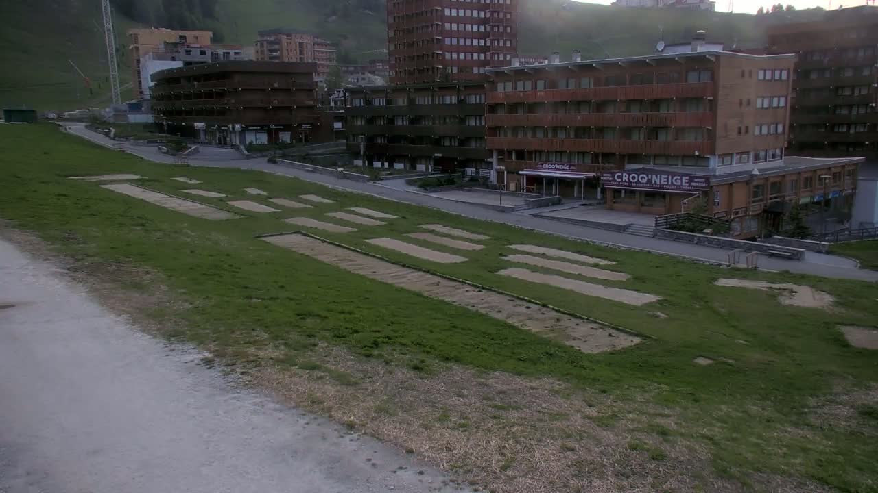 A grassy field with rectangular patches of bare earth sits below several multi-story buildings on a hillside, under an overcast sky.