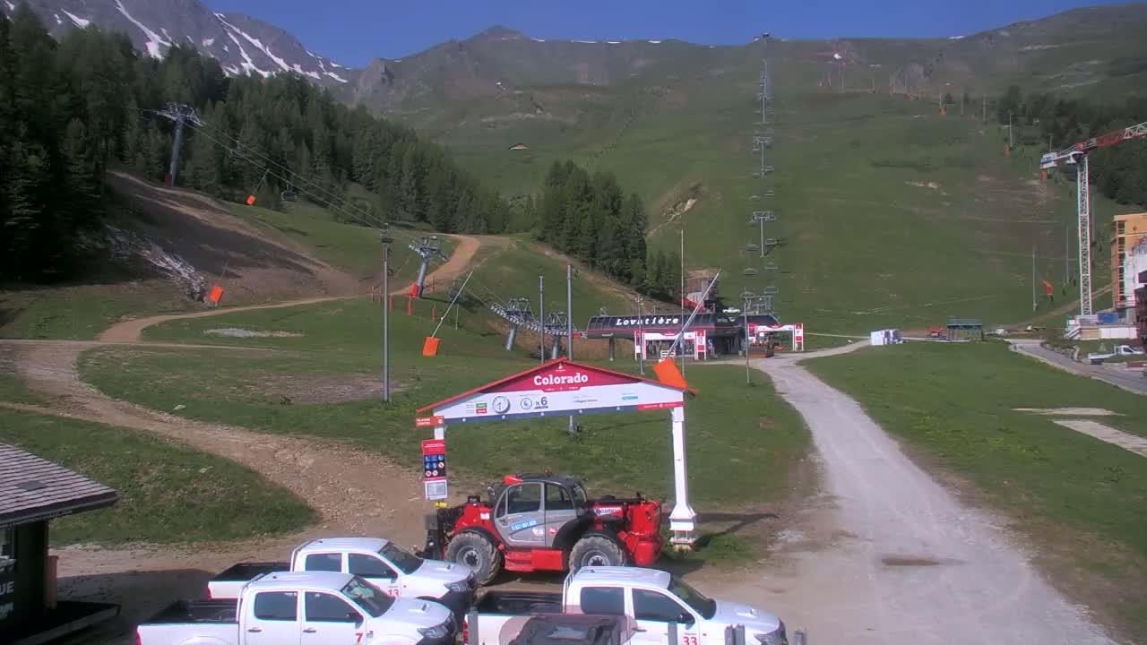A sunny mountainside ski resort scene shows several chairlifts, a small building, a few parked vehicles, and a construction crane in the distance.