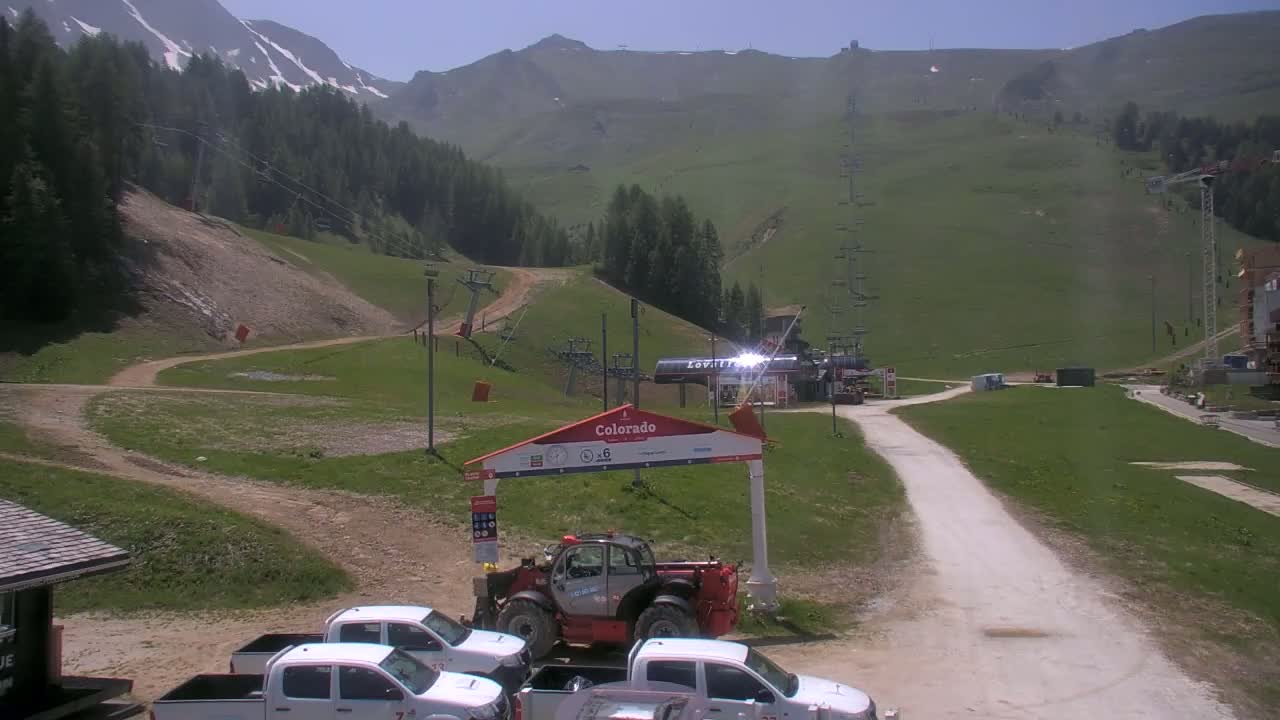 A sunny day at a mountain resort shows a grassy hillside with ski lifts, a dirt road, and several parked pickup trucks near a small building.