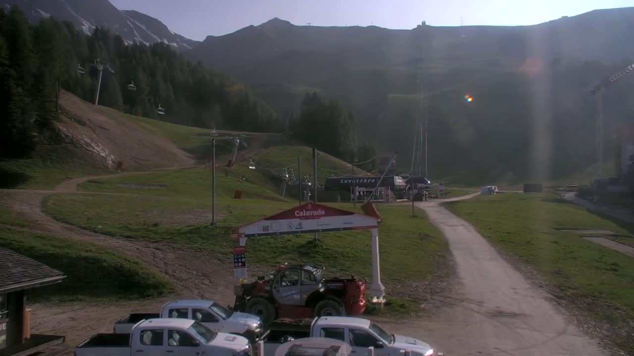 A sunny, hazy day at a ski resort shows a grassy area with several utility vehicles parked near a ski lift and a mountain in the background.