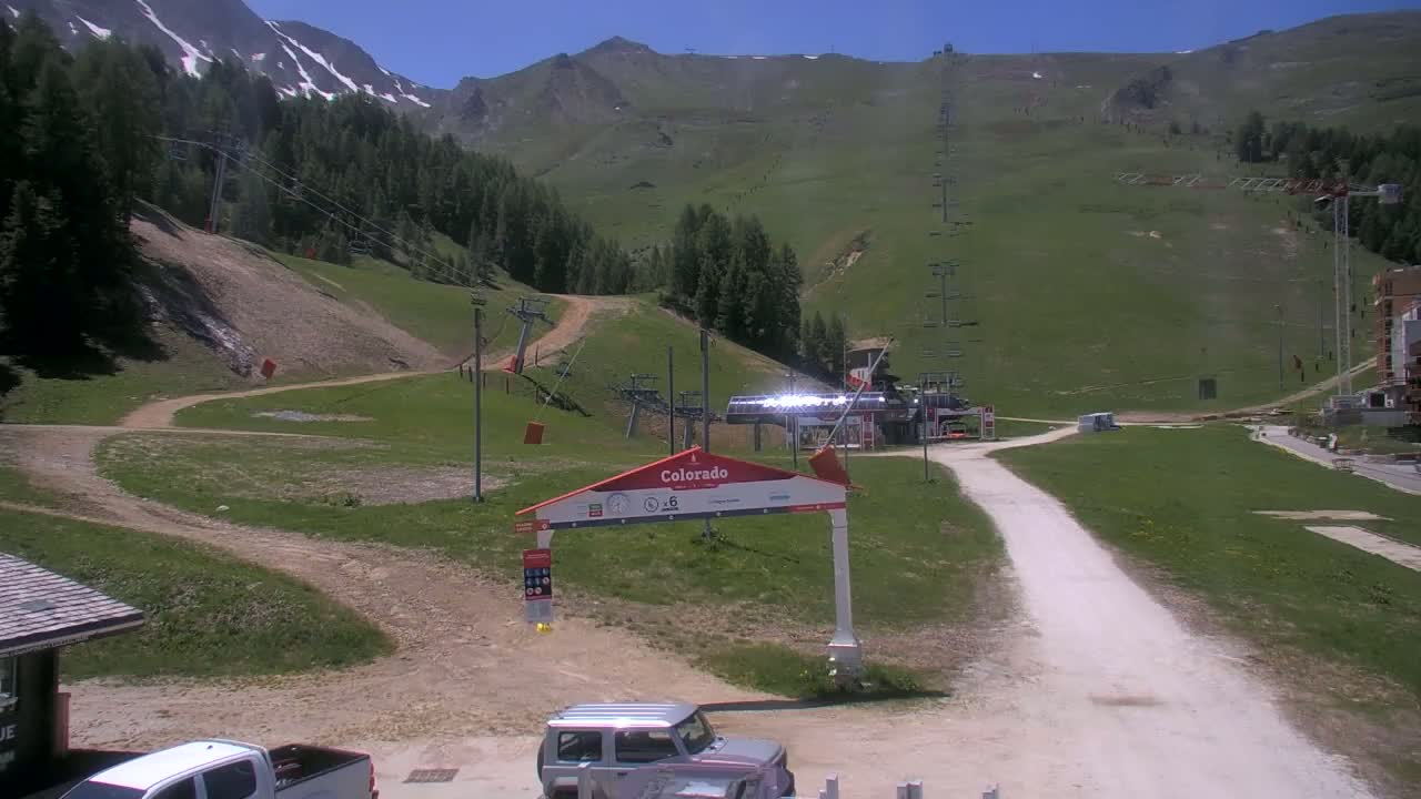 A sunny day at a mountain ski resort with chairlifts, grassy slopes, and a dirt road leading to a base area with a few vehicles parked.