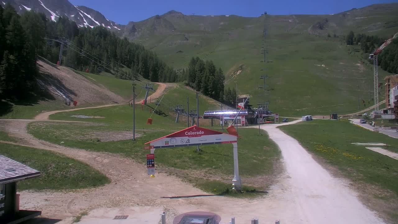 A sunny view of a mountainside ski resort in the summertime, showing grassy slopes, chairlift infrastructure, and a light-colored dirt road leading to a base area.