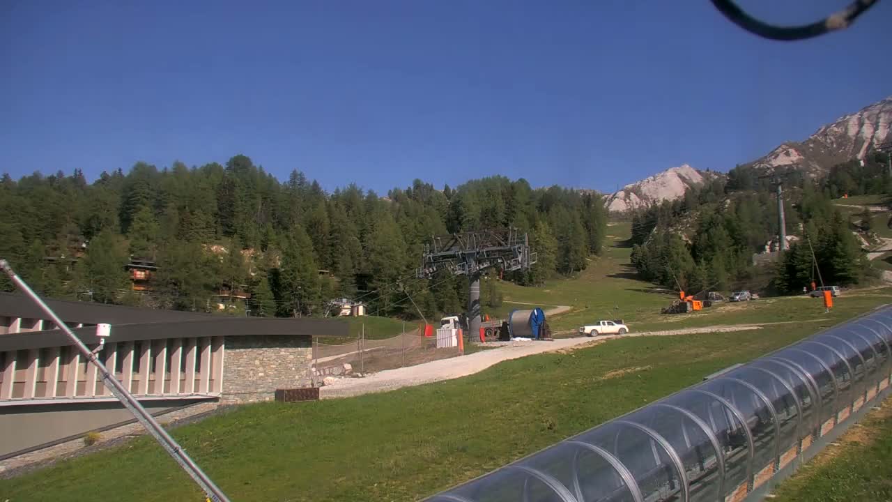 A ski lift base station is visible in a sunny, mountainous area with green grass and coniferous trees, alongside a covered, enclosed lift.