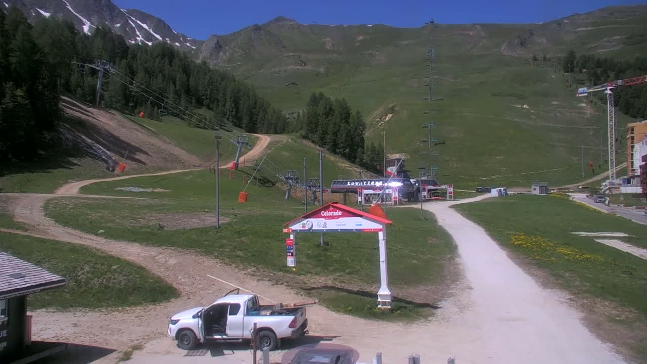 A sunny day at a ski resort shows a mountainside with chairlifts, grassy areas, and a dirt road leading to a building; a white pickup truck is parked near a sign.