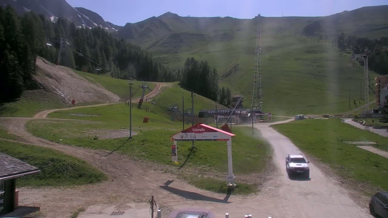 A sunny, dusty mountainside scene shows a dirt road leading to a ski lift and a few buildings, with a silver SUV driving down the road.