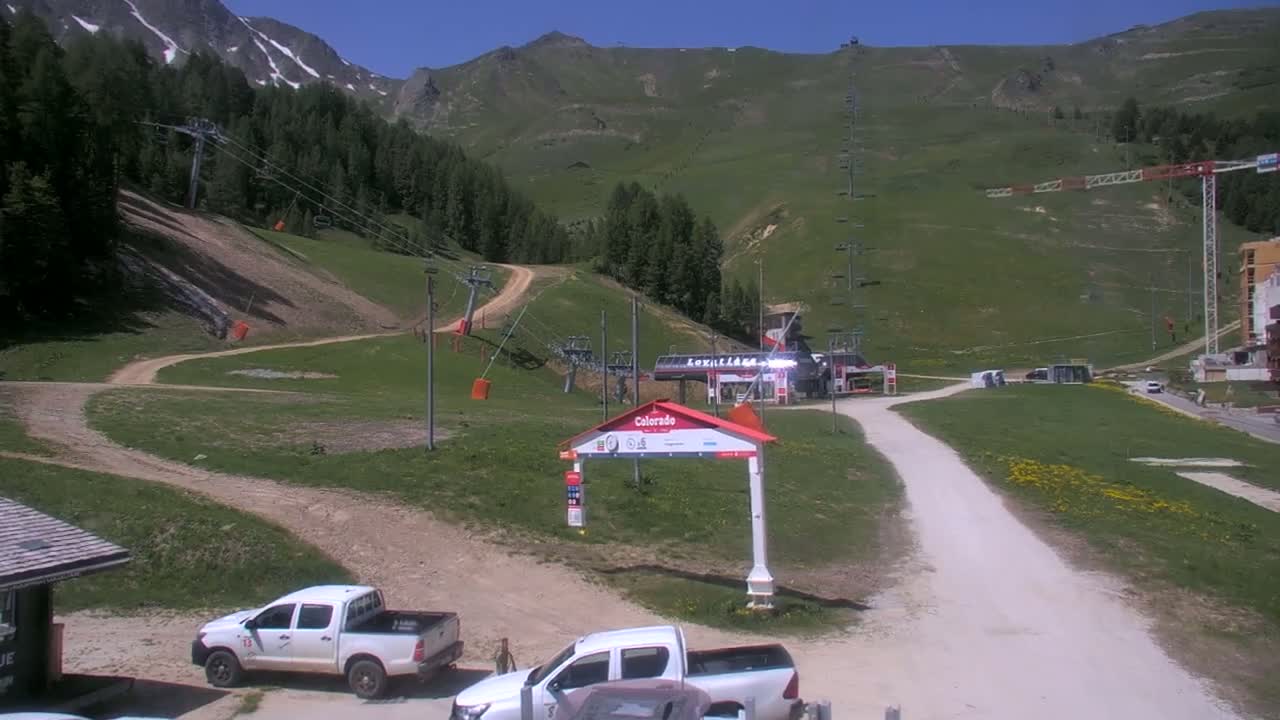A sunny mountainside ski resort shows a light-colored dirt road leading to a ski lift base, with several parked pickup trucks and a construction crane in the background.