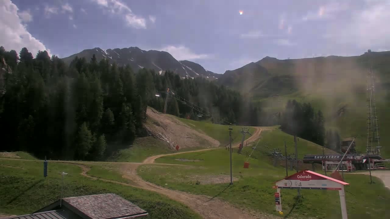 A partly cloudy day reveals a mountain ski resort with chairlifts, grassy hills, and a dirt road winding through the landscape, with hazy conditions in the distance.