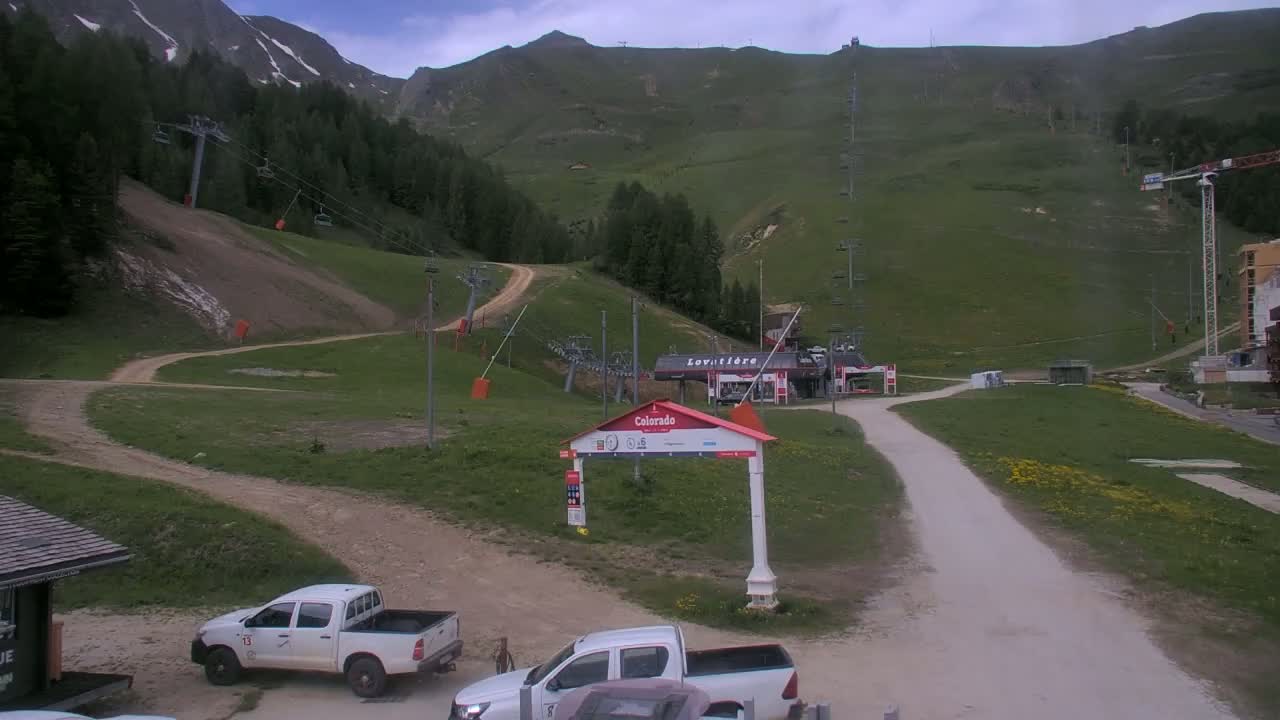 A mostly cloudy day at a ski resort shows several chairlifts, grassy hills, dirt roads, and two parked pickup trucks.