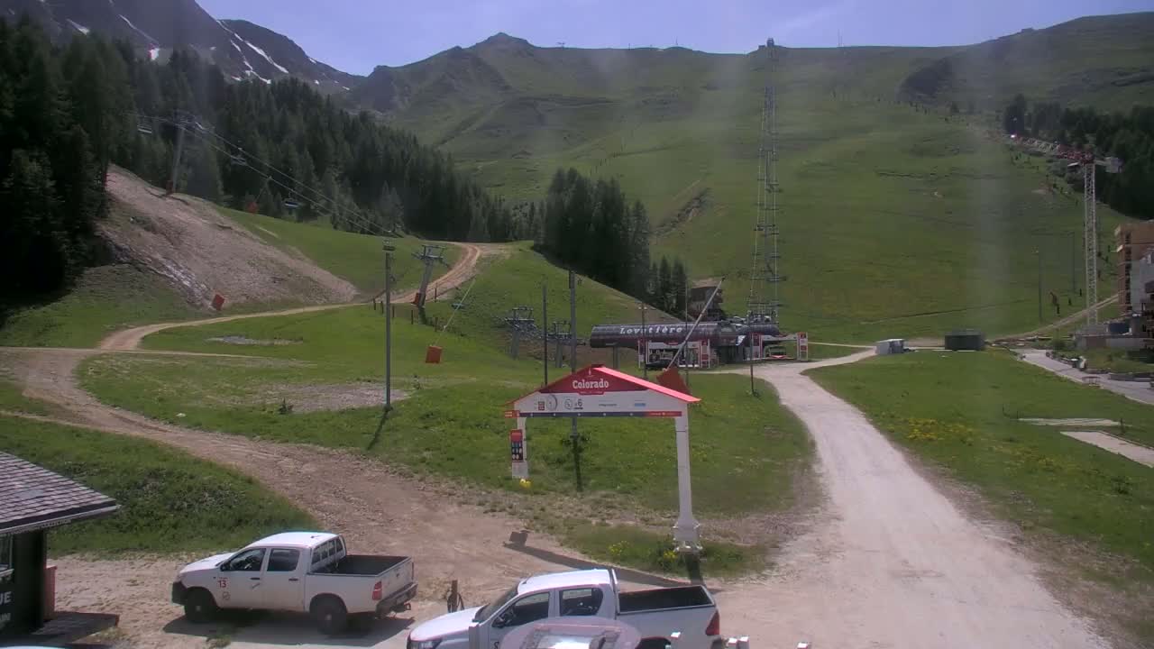 A sunny, green mountainside with a dirt road leading to a ski lift base area where two pickup trucks are parked.