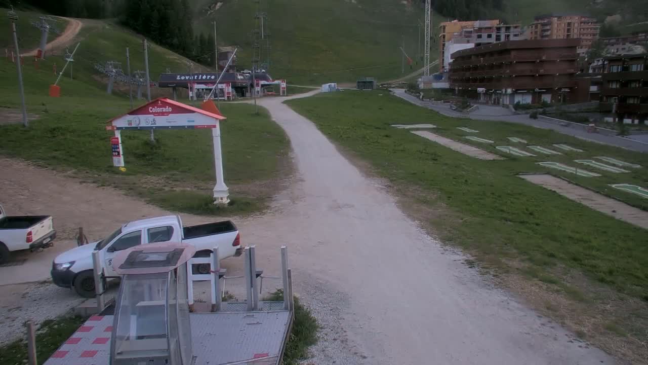 A sunny, green mountainside with a dirt road leading to a ski lift base area where two pickup trucks are parked.