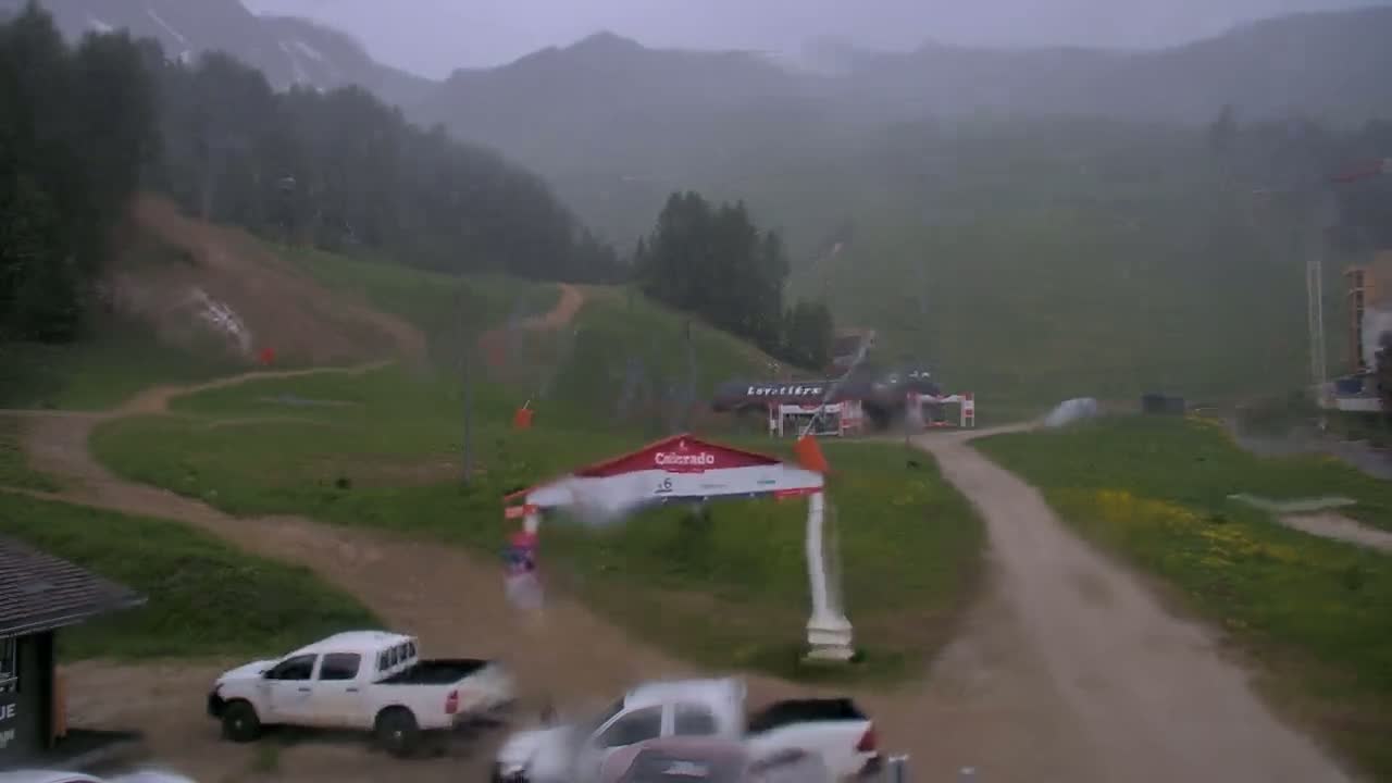 A rain-soaked mountainside with dirt roads, a few parked trucks, and a building in the distance under heavy rainfall.