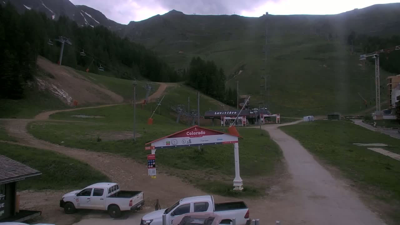 A mountainside ski resort scene under an overcast sky shows two white pickup trucks parked near a gateway, with ski lifts and grassy hills in the background.
