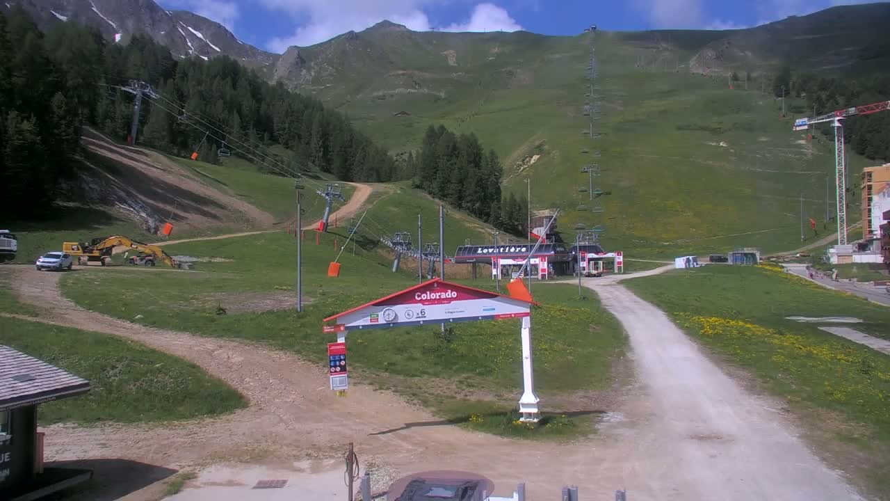 A sunny day at a mountain ski resort shows construction equipment near a lift station, with a grassy hillside and mountain range in the background.