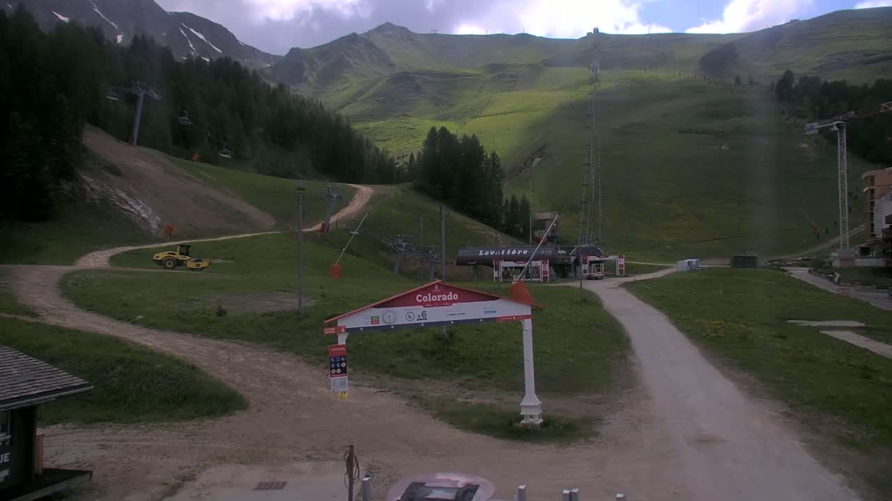 A mostly cloudy day reveals a mountain ski resort with grassy slopes, chairlifts, and a small yellow piece of construction equipment parked on a dirt road.