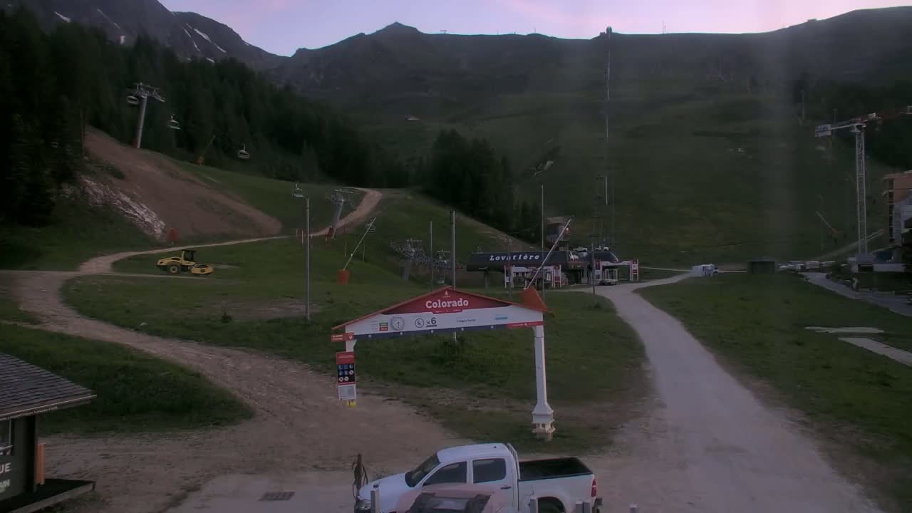 A mostly clear evening sky overlooks a ski resort with several chairlifts, a dirt road, and a white pickup truck parked near a gateway.