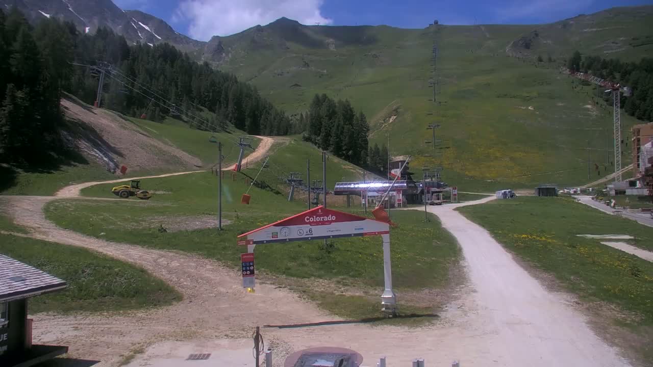 A sunny day at a mountain ski resort shows a grassy hillside with ski lifts, a road, and a piece of construction equipment.