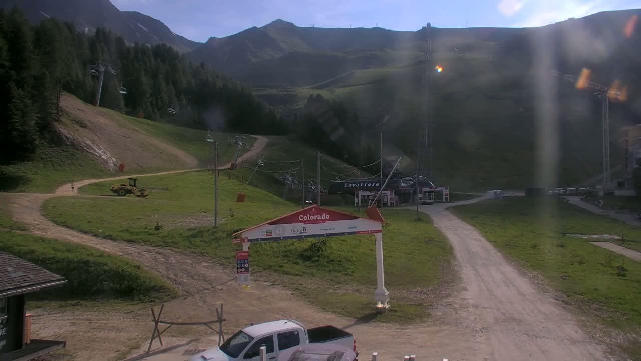 A mountainside view on a partly sunny day shows a dirt road leading to a ski lift base area with a white pickup truck parked nearby, a yellow road roller on a grassy area, and a mountain range in the background.
