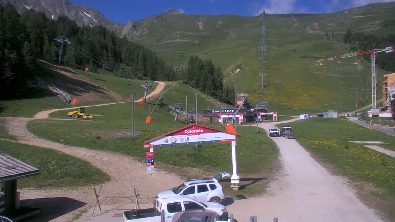 A sunny day at a ski resort shows several vehicles parked near a lift station and a small piece of construction equipment on a hillside with chairlifts and green grass.