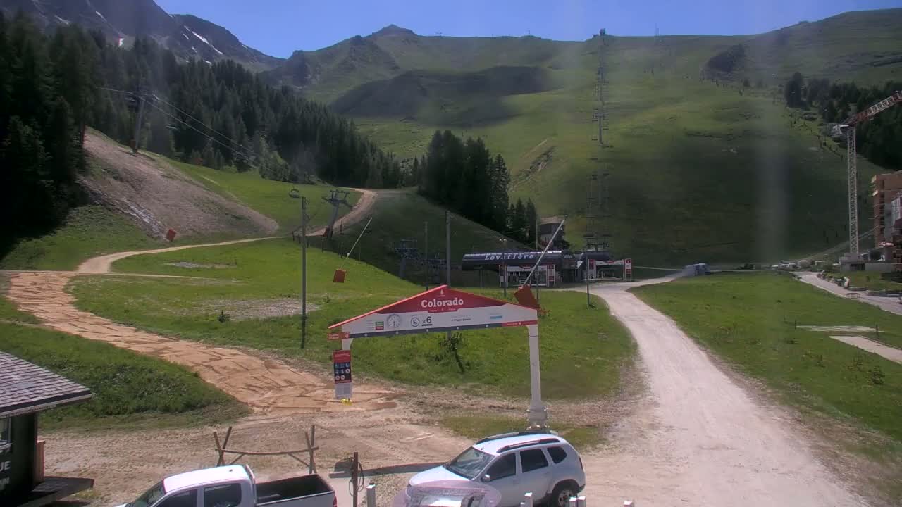 A sunny day at a mountain resort shows a grassy hillside with ski lifts and a dirt road leading to a parking area with several vehicles.