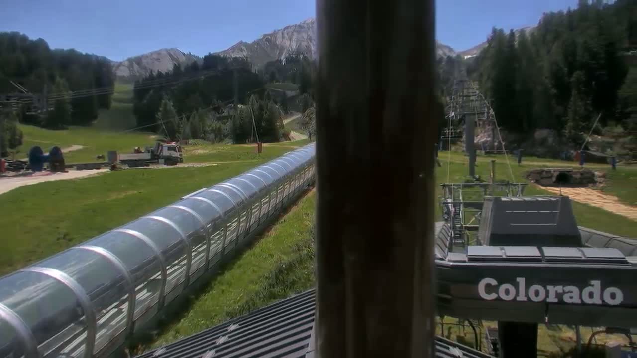 A long, covered conveyor belt extends across a grassy area below a chairlift in a mountainous region under sunny skies.