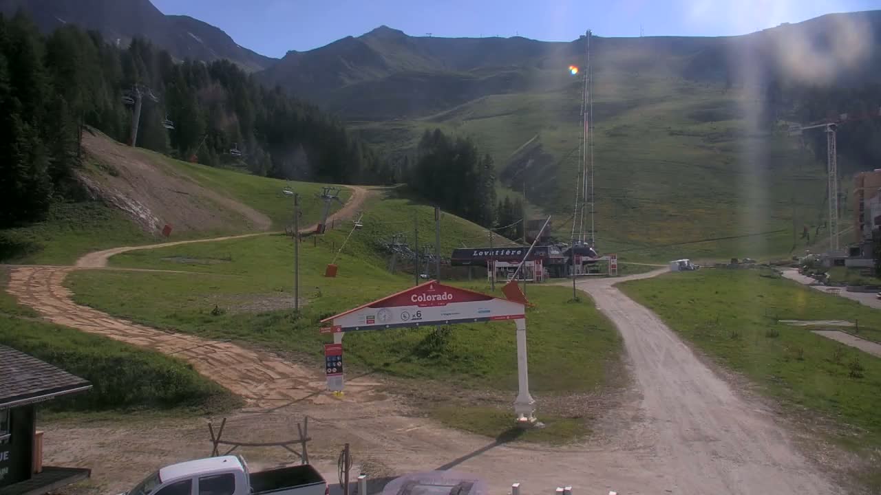 A sunny day at a mountain resort shows grassy hills, chairlifts, and a dirt road leading to a building.