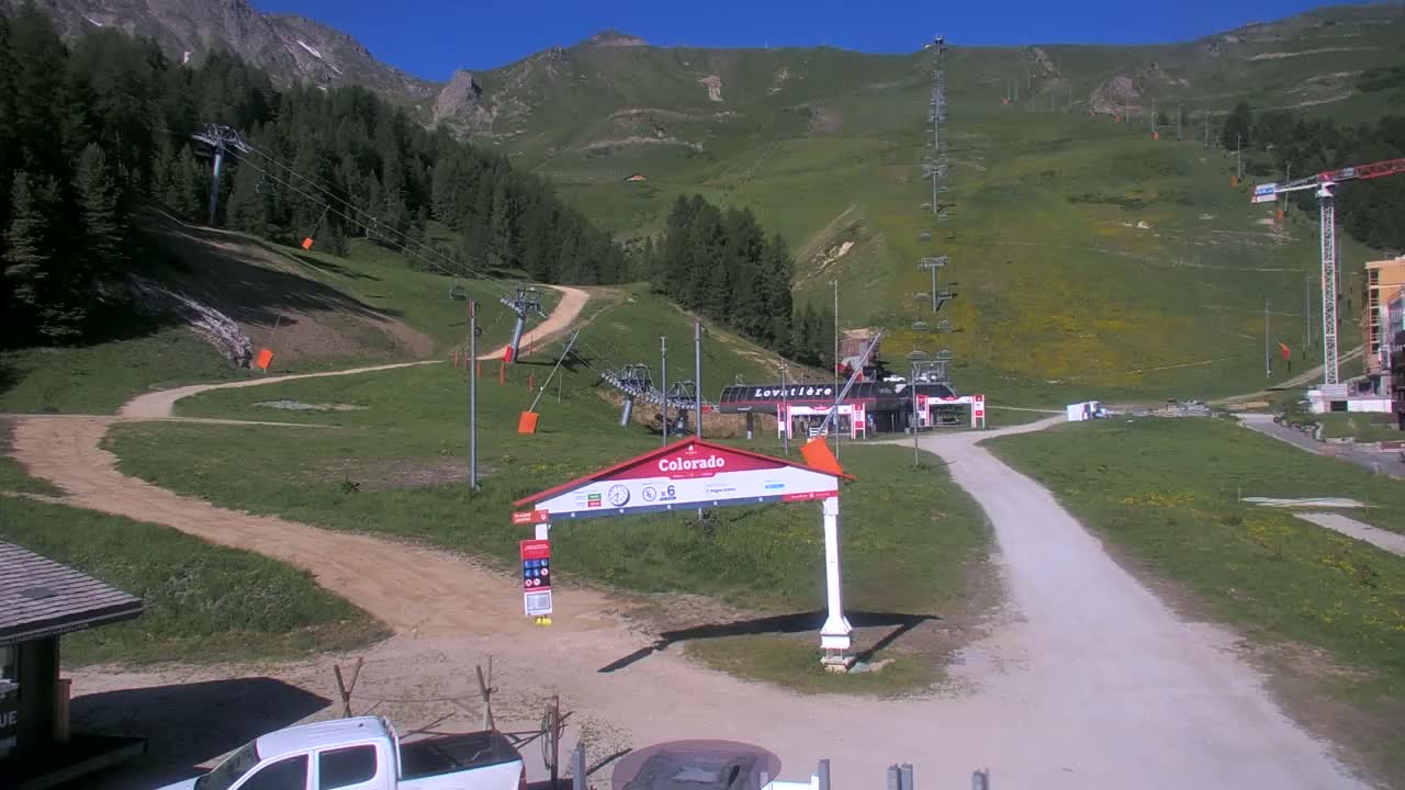 A mountainside ski resort in sunny conditions shows grassy slopes, chairlifts, and a dirt road leading to a lift station.