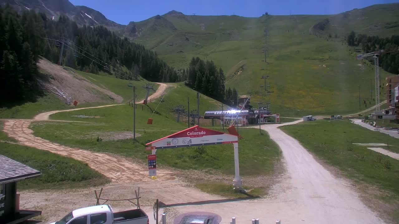 A sunny view of a mountain ski resort in summer, showing grassy slopes, dirt roads, ski lifts, and a few vehicles.