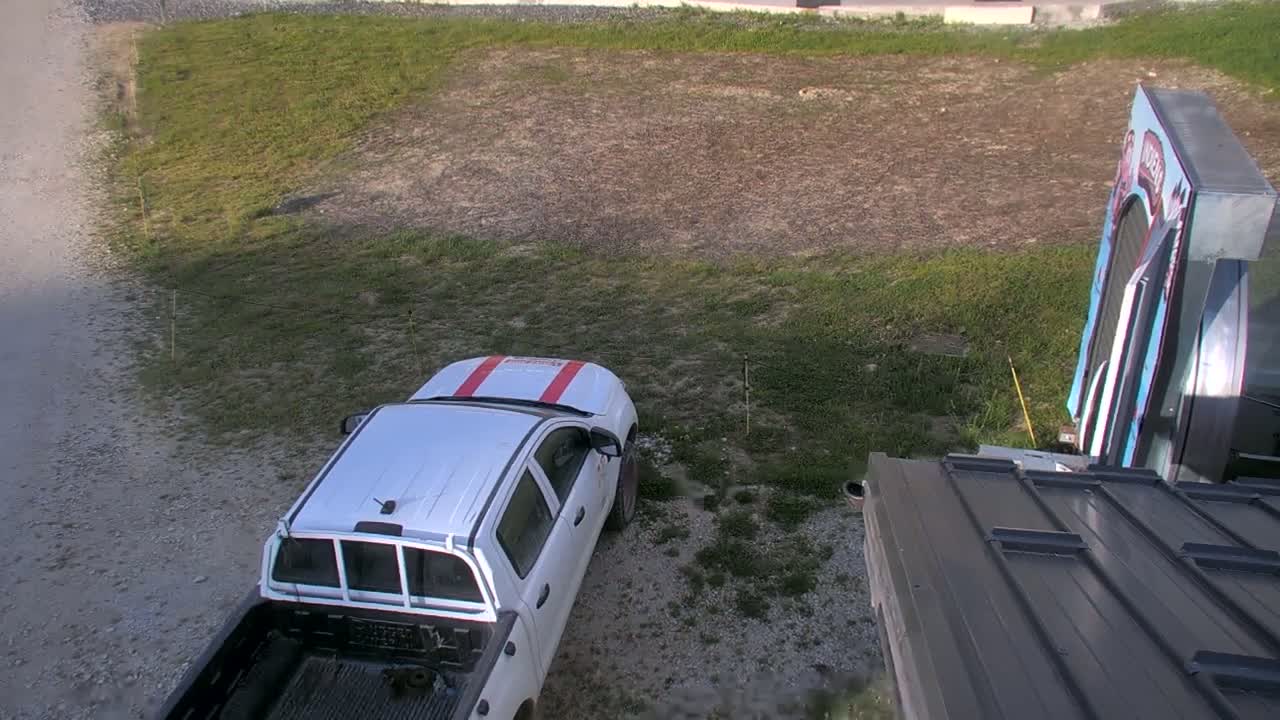 A white pickup truck is parked in a gravel area next to a patch of grass and a dark-colored metal roof.