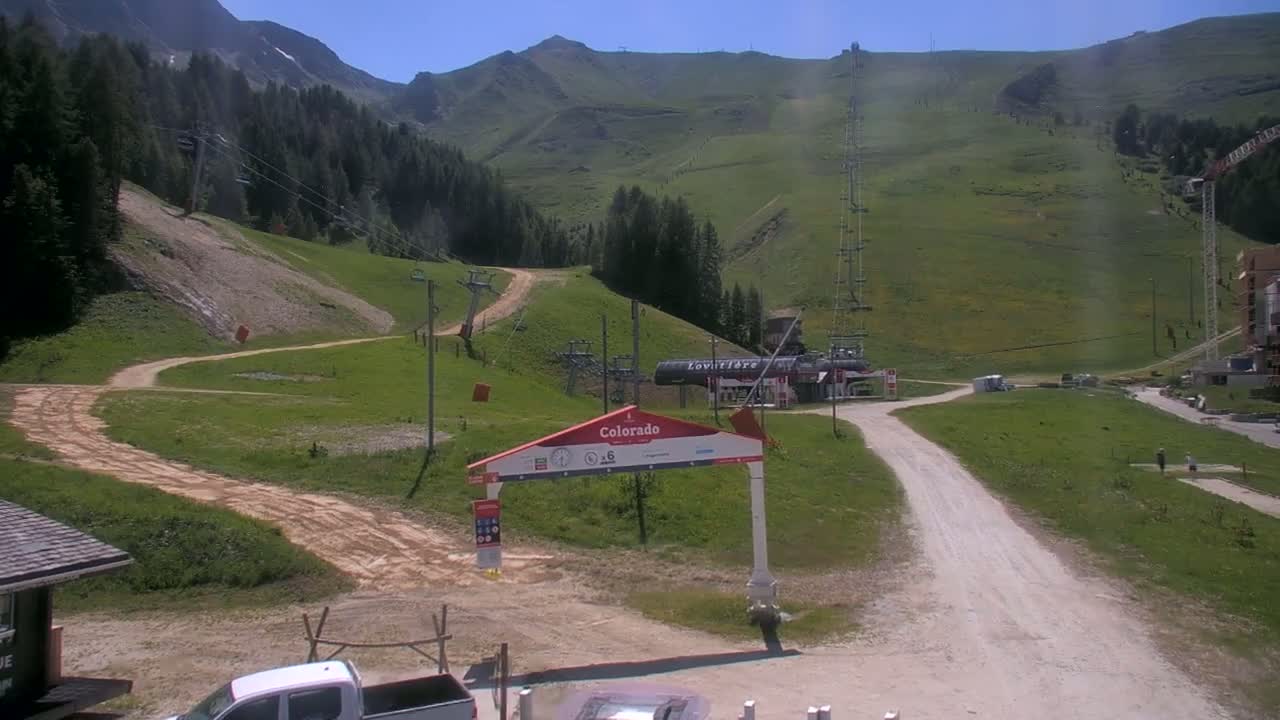 A sunny mountain scene shows a dirt road winding through green grassy hills with ski lifts and a few buildings in the distance.