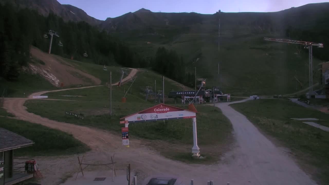 A mountain ski resort at dusk with chairlifts, dirt roads, and a construction crane visible on a hillside.